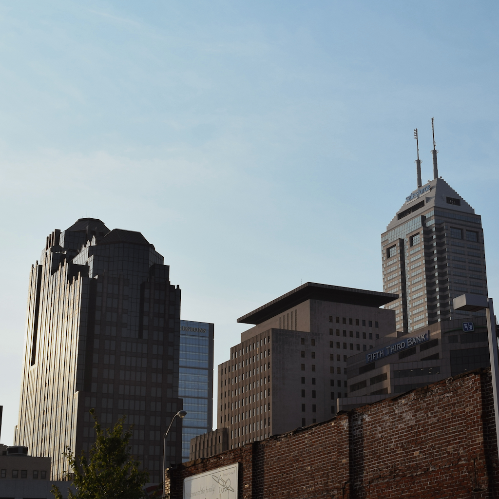 A low-angle photograph of a city skyline featuring the historic South Bank Tower and other mid-century skyscrapers against a pale blue sky, with a brick wall in the foreground.